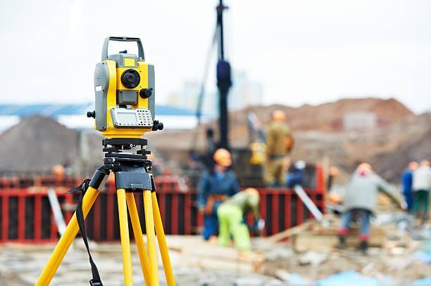 A yellow theodolite on a tripod surveys a construction site with workers, blurred in the background