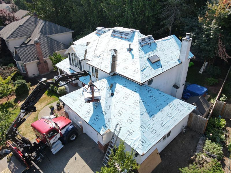 Aerial view of house roof being worked on, crane lifting materials. Blue underlayment visible.