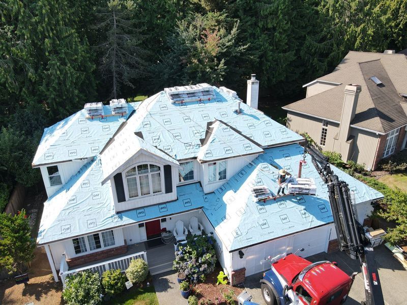 House roof being re-shingled; workmen on roof with crane. Bright blue underlayment visible. Red truck below.