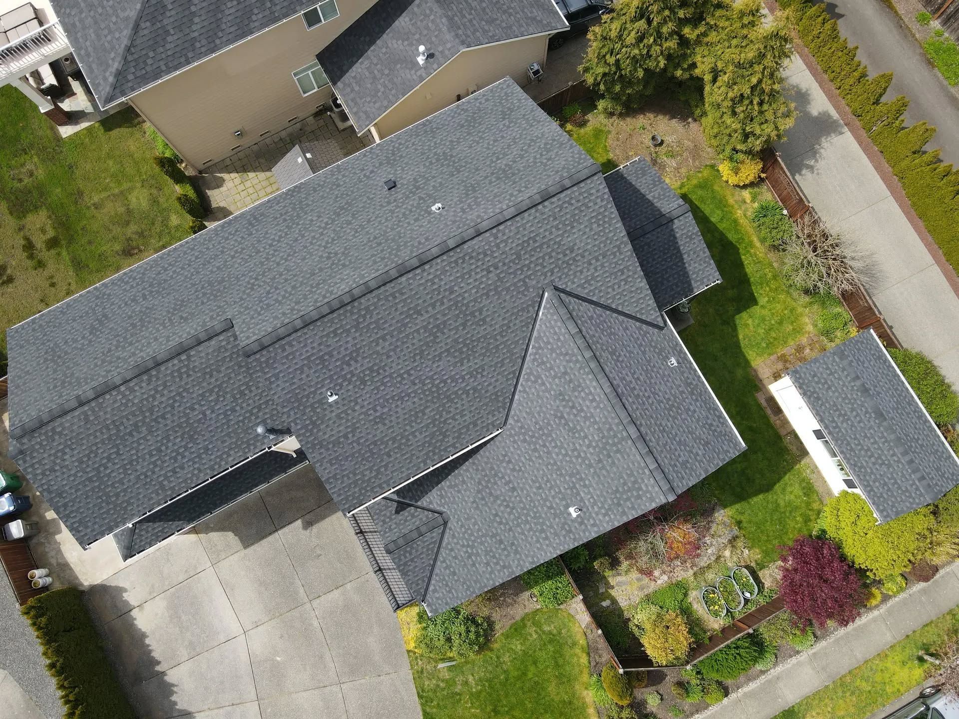 Overhead view of a house with a dark gray shingle roof, surrounded by green grass and a small garden.