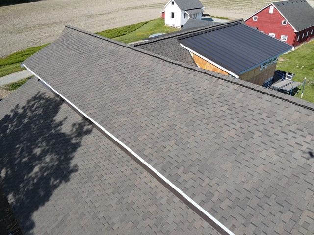 Overhead view of a roof with brown shingles and white trim, overlooking buildings and a field.