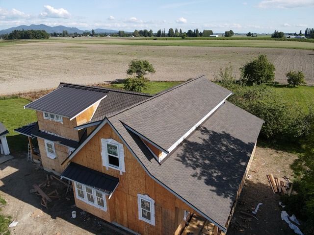 Partially-built house with light-brown siding and a dark roof, in a rural setting with fields and trees.