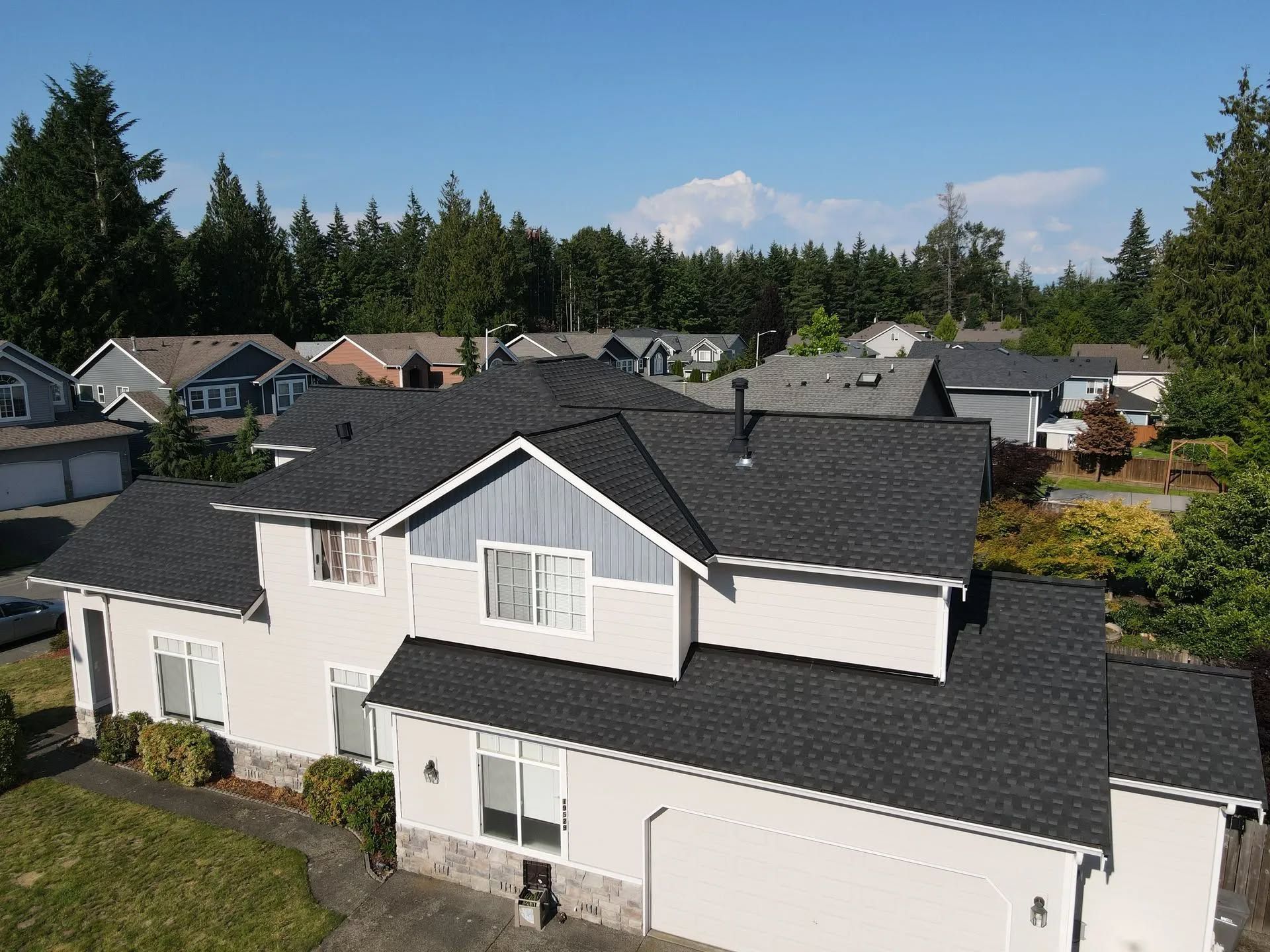 Two-story house with dark roof and blue trim, trees and mountain in the background under blue sky.