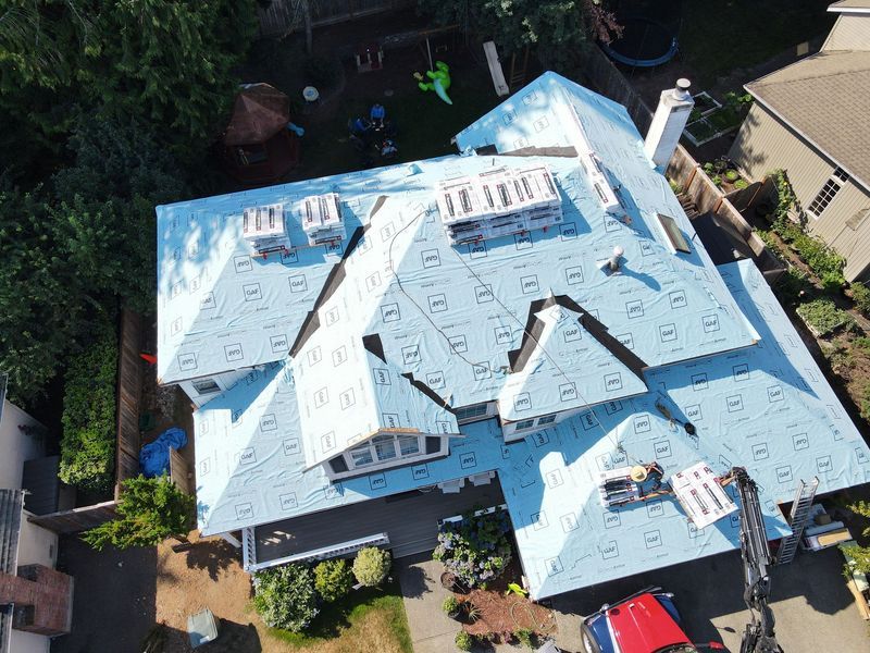 Aerial view of house roof being renovated, blue underlayment visible, construction in progress.