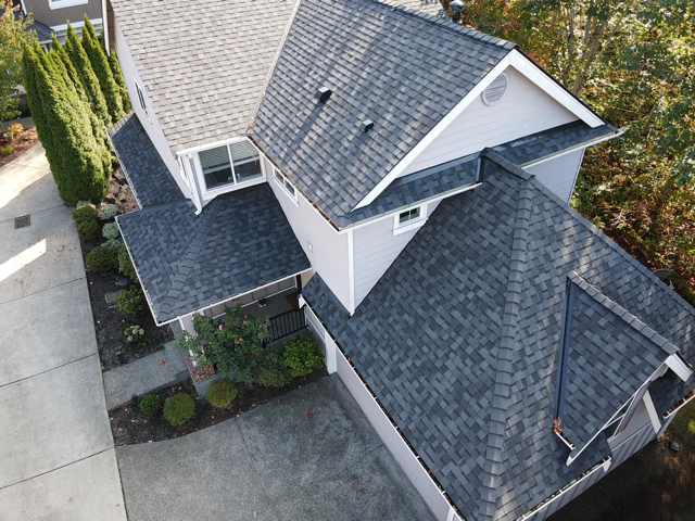 Overhead view of a gray-roofed two-story house with a driveway, surrounded by trees and landscaping.