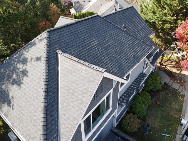 Dark gray shingled roof of a house, seen from above, with trees and lawn.