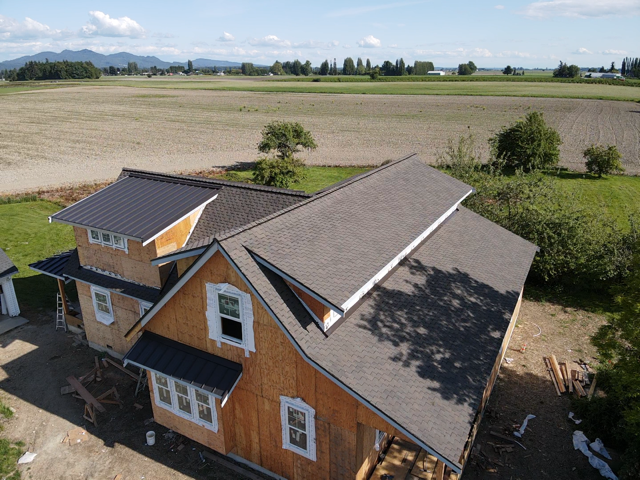 House under construction with two-tone roof: black metal and gray shingles, set in a field.