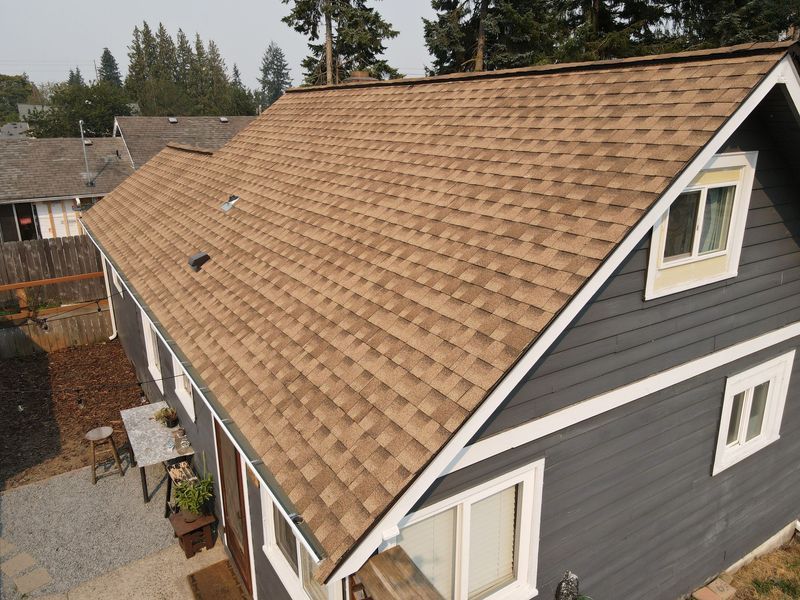 A house with a brown shingled roof, gray siding, and white window frames.