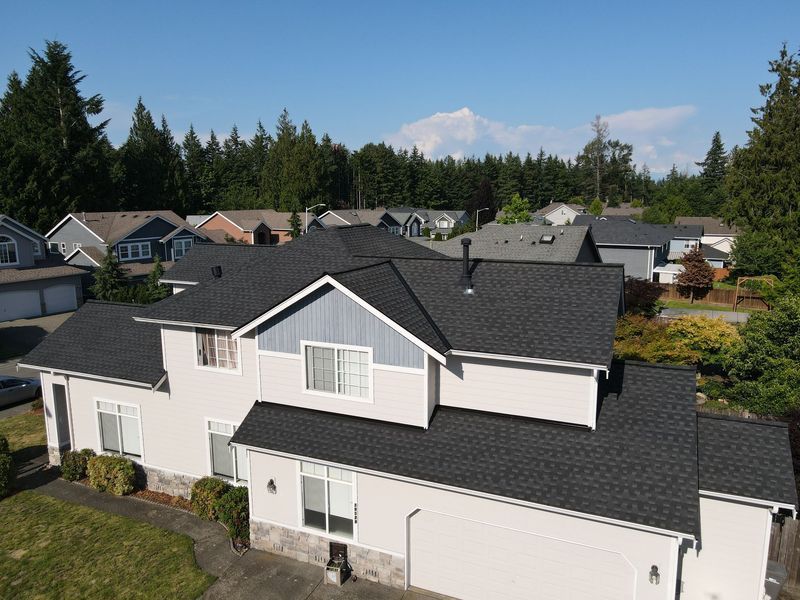 Two-story house with dark gray roof, light gray siding, and blue accents. Residential neighborhood with trees and a mountain in the background.