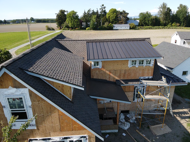 House under construction with contrasting shingle and metal roof sections, wood siding, and scaffolding.