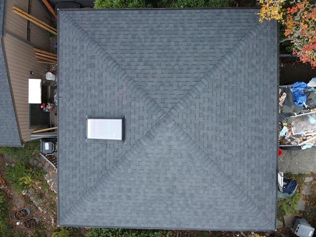 Overhead view of a gray shingled roof with a rectangular vent. Surrounding are trees and a building.