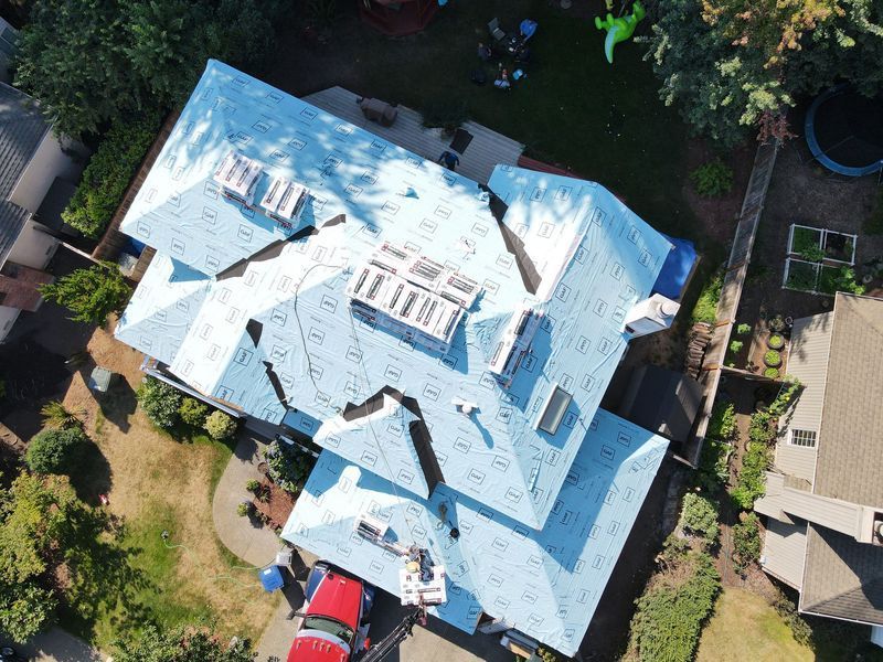 Aerial view of a blue-roofed house during roofing work, with skylights and surrounding yard.