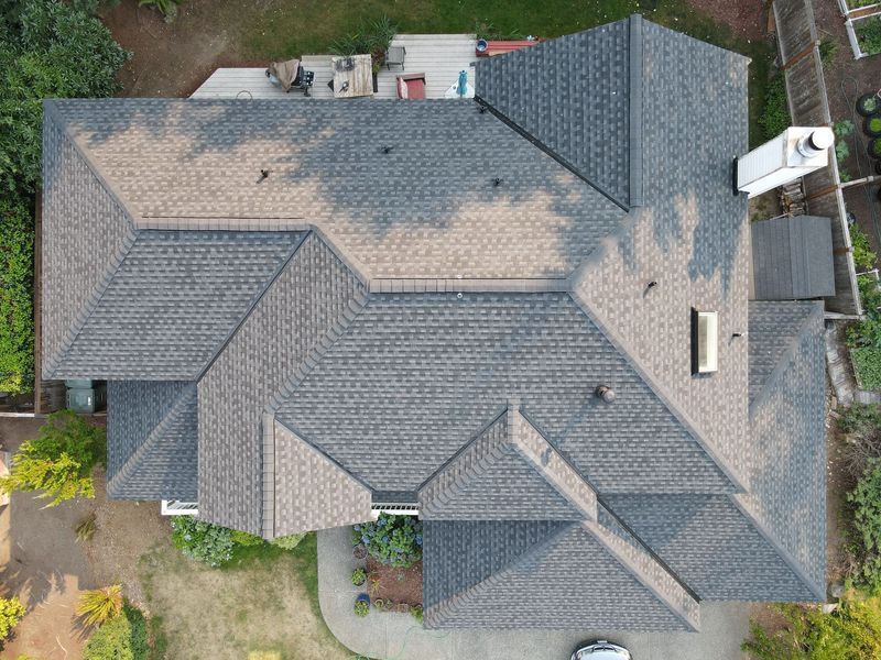 Overhead view of a house with a gray shingle roof, surrounded by green trees and a white propane tank.