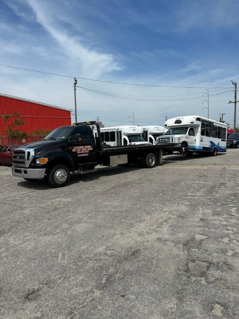 Black tow truck towing a white and blue bus, parked in a lot under a blue sky.