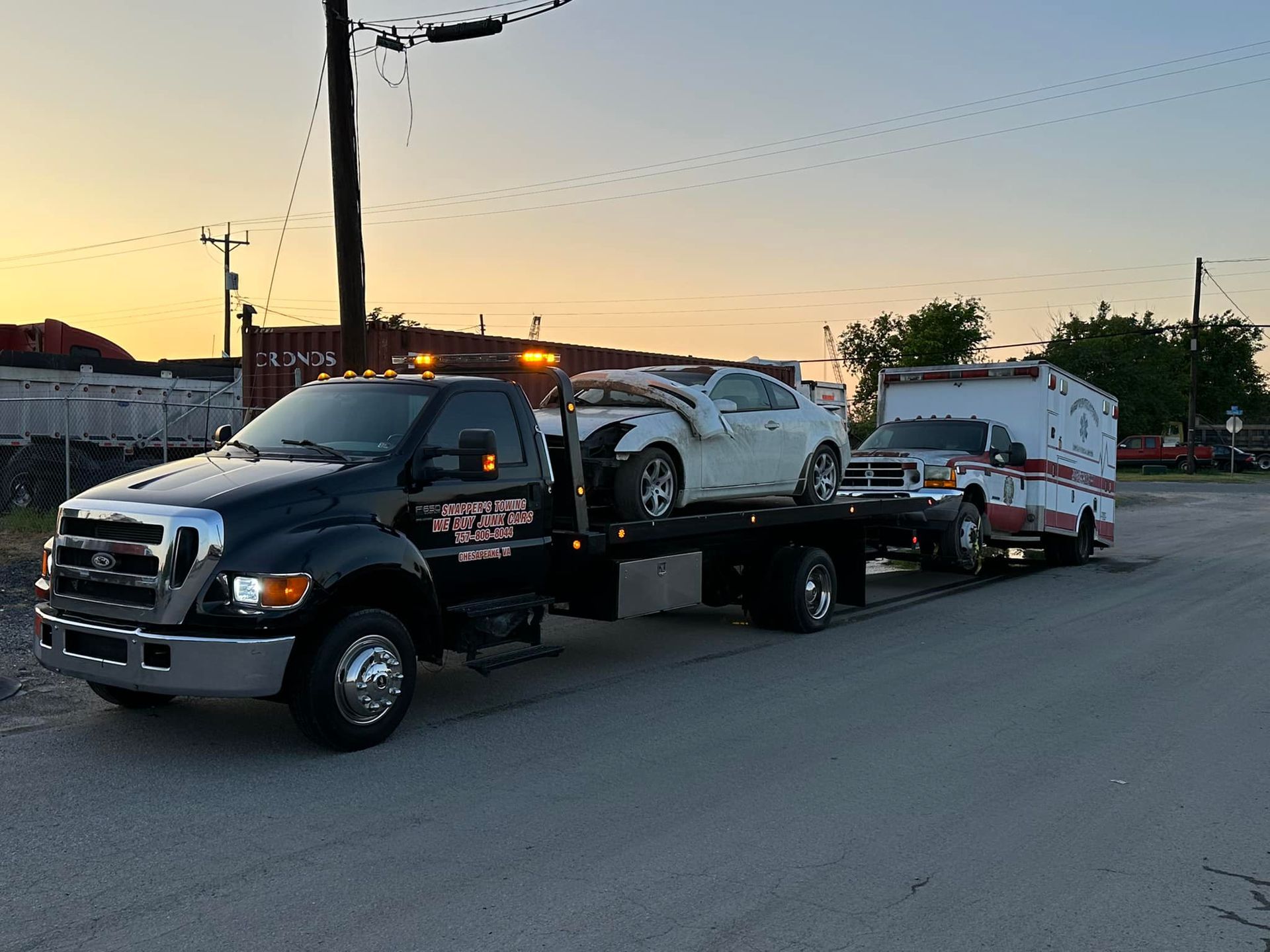 Black tow truck with damaged white car and ambulance, transporting them on a city street at dusk.