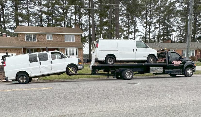 A white van is being towed on a flatbed truck in front of a house.