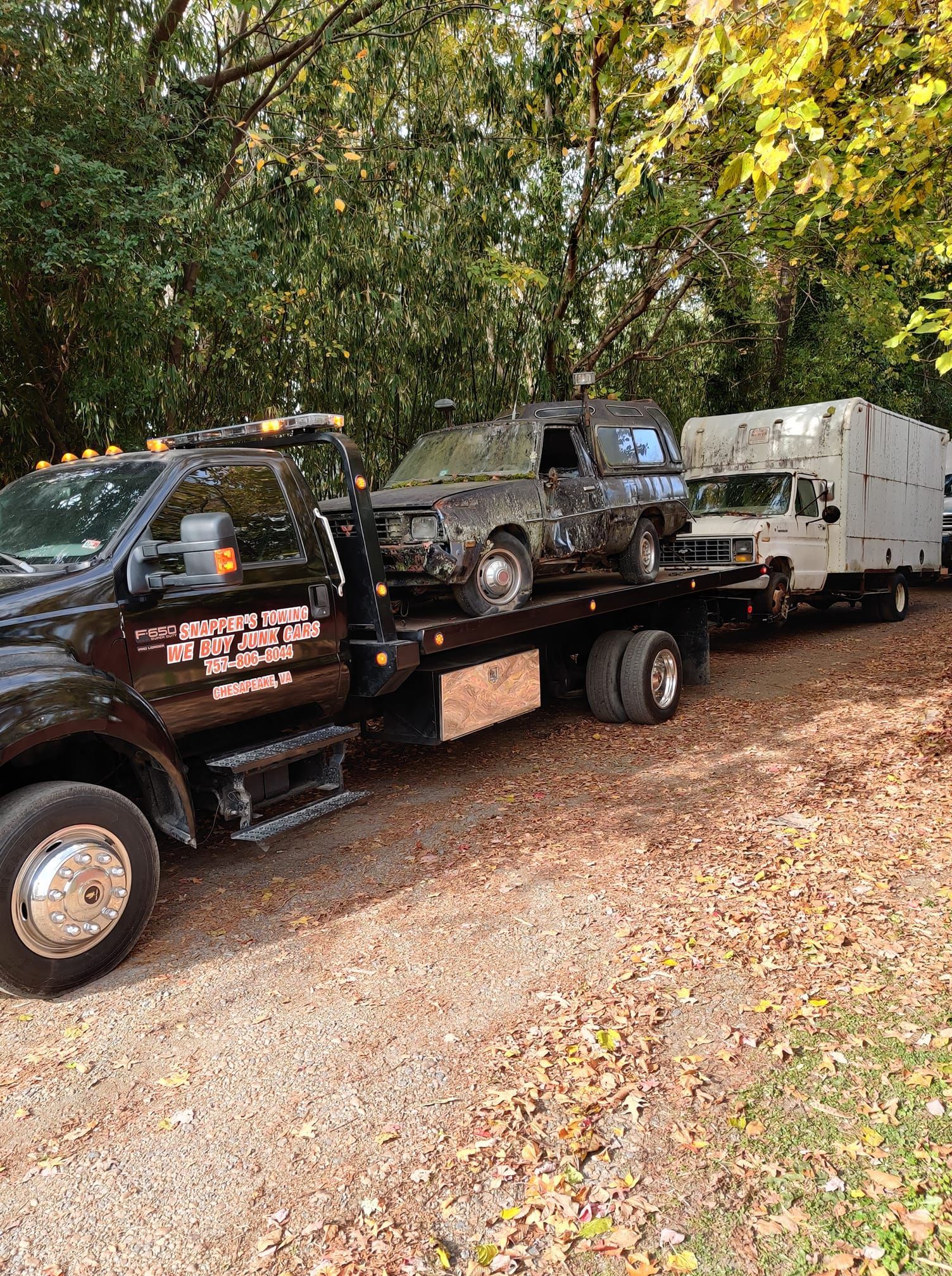Tow truck transporting a damaged vehicle and a large white box truck on a gravel road. Autumn setting with trees.