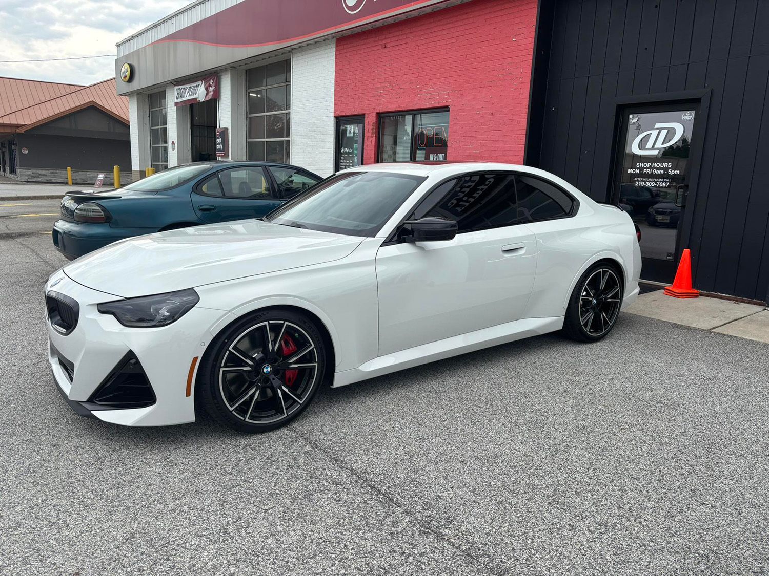 White BMW coupe parked in front of a building with red and black exterior, black wheels, and tinted windows.