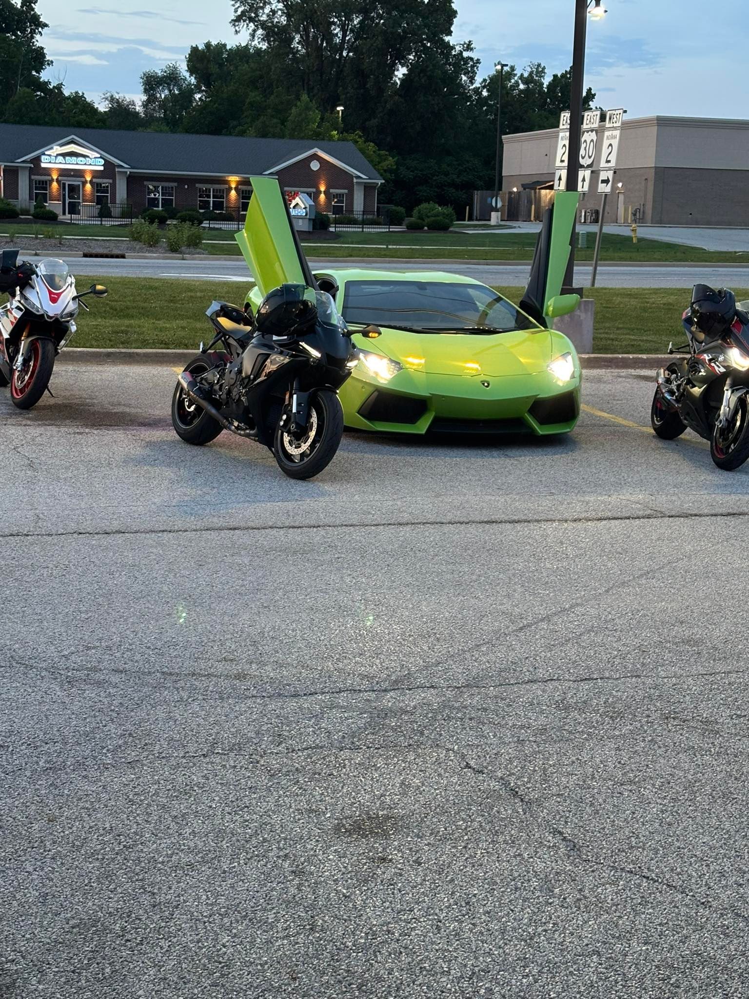 A lime green Lamborghini with butterfly doors open, flanked by black motorcycles in a parking lot.
