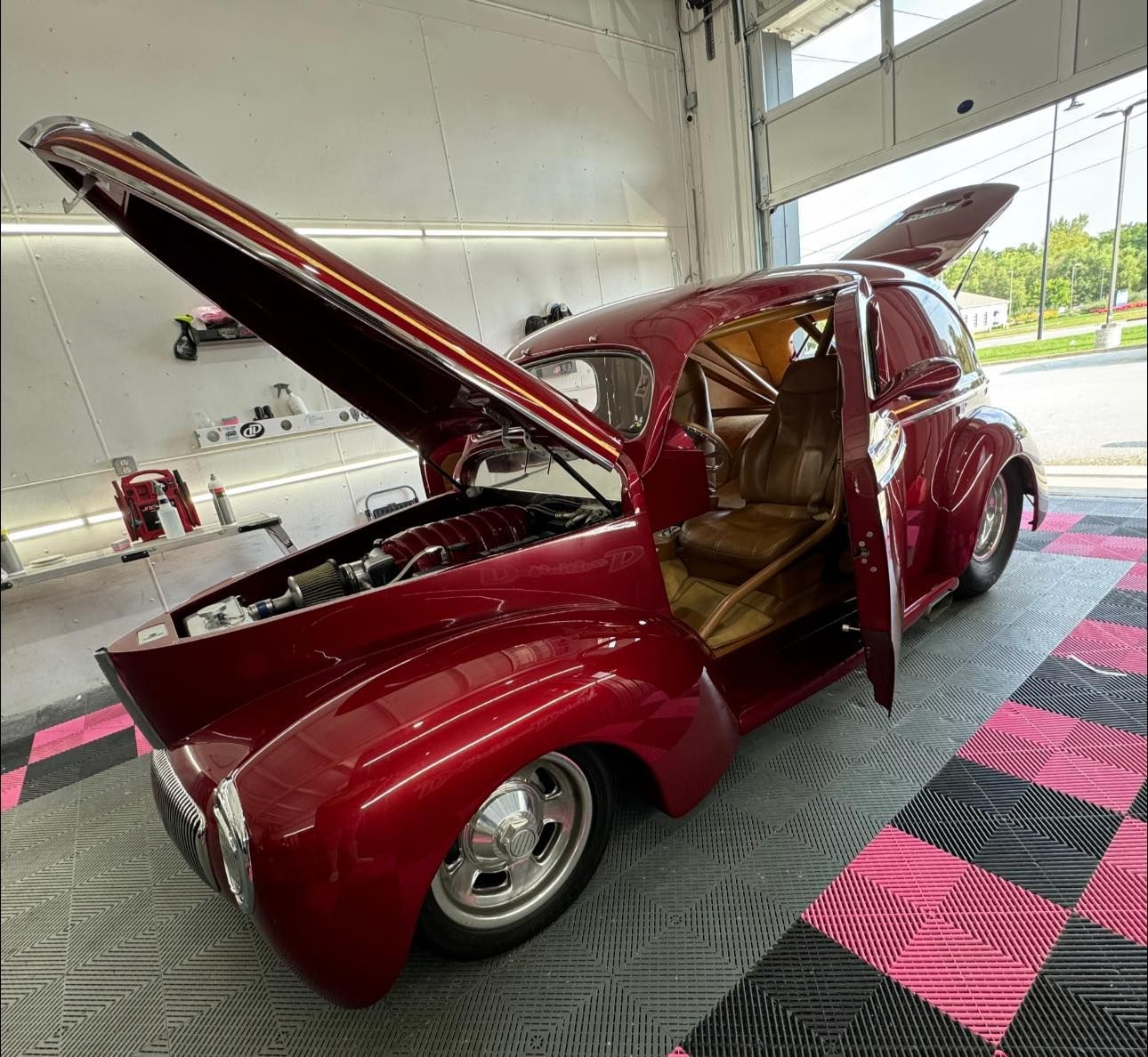 A maroon custom hot rod with open doors and hood, parked on a patterned garage floor.