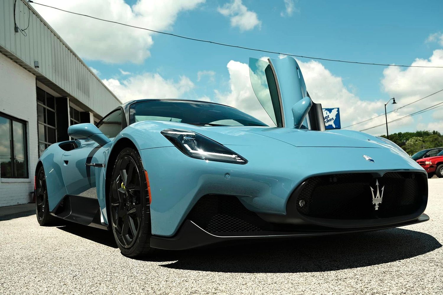 Light blue Maserati MC20 sports car with open butterfly doors, parked in front of a building on a sunny day.
