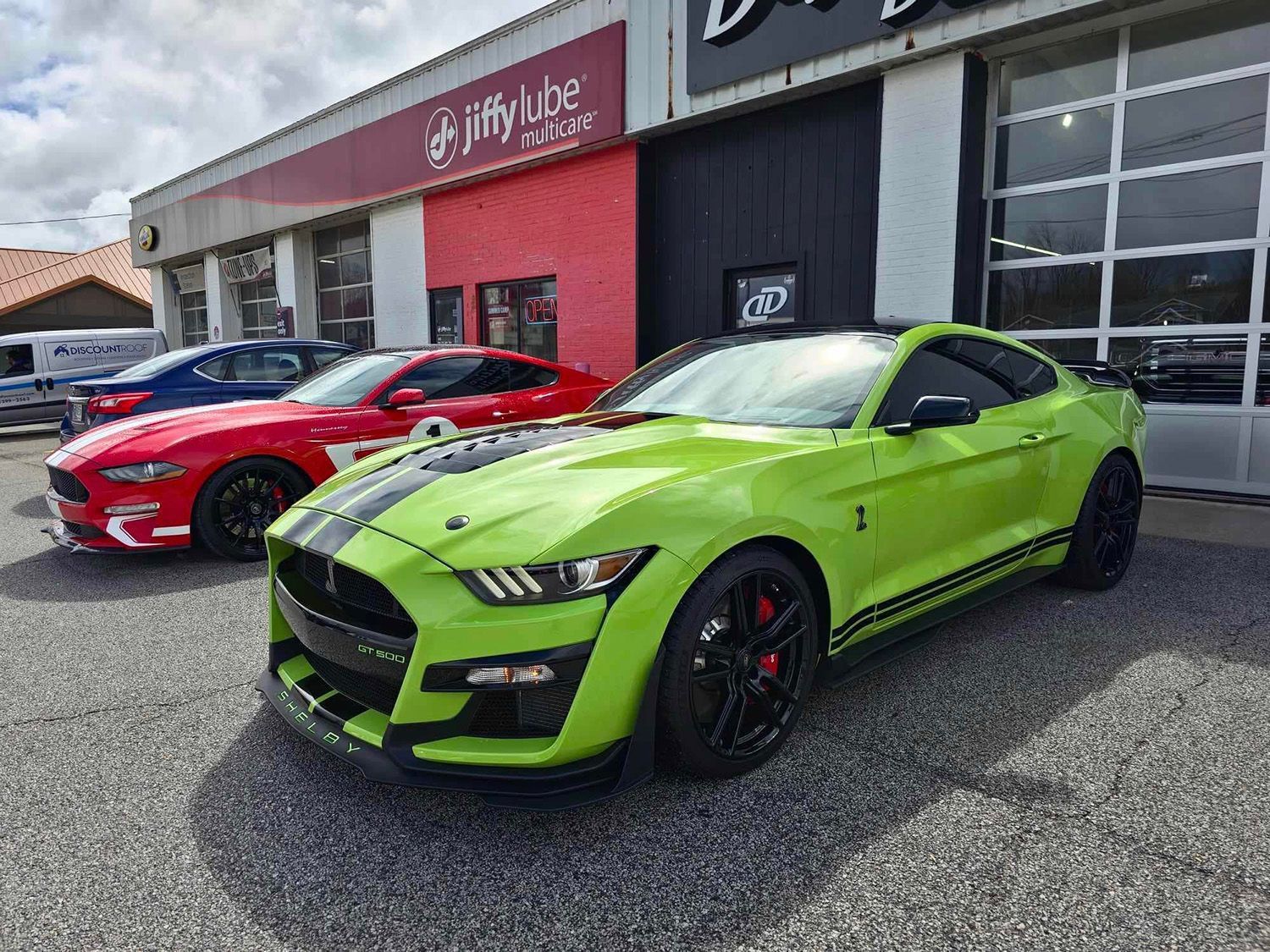 Green Ford Mustang Shelby in front of a Jiffy Lube, parked next to a red Mustang.
