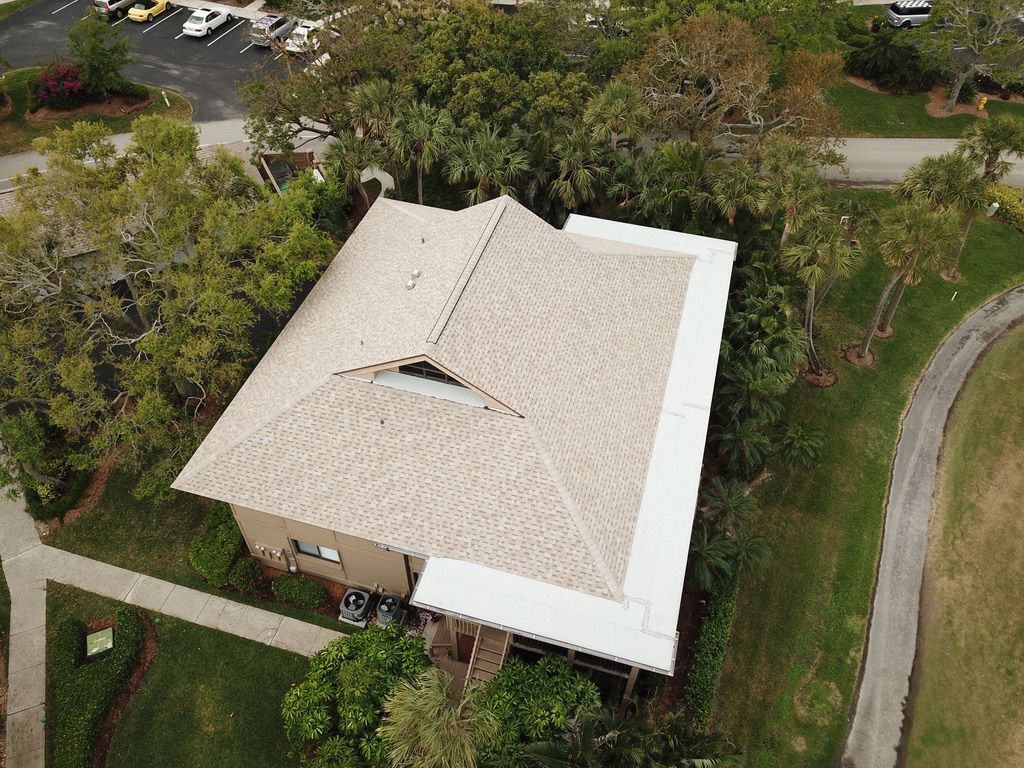 Aerial view of a light-colored house with a white roof surrounded by trees and a curved driveway.