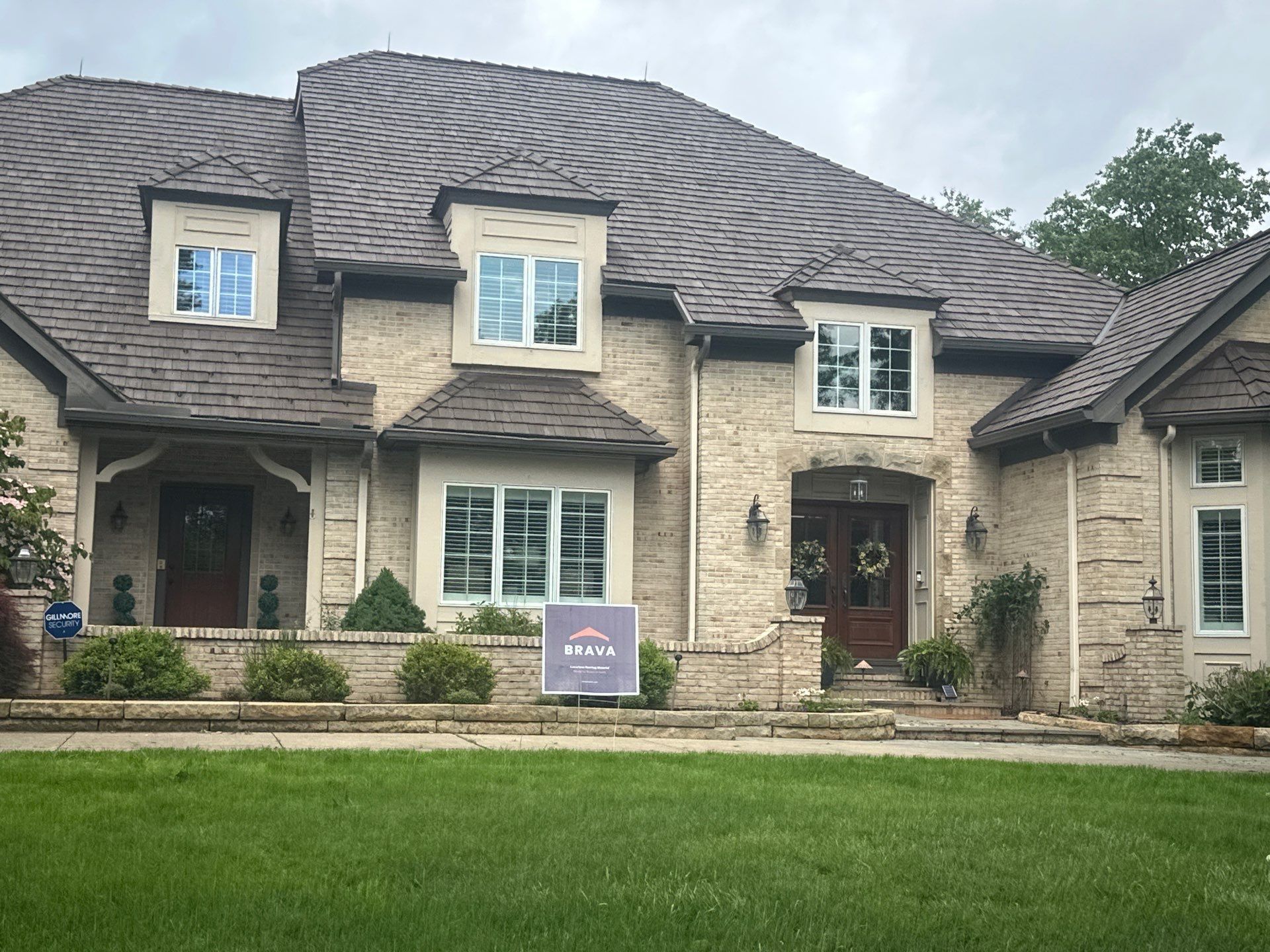 Stone suburban house with gray shingled roof, manicured lawn, and small sign in front