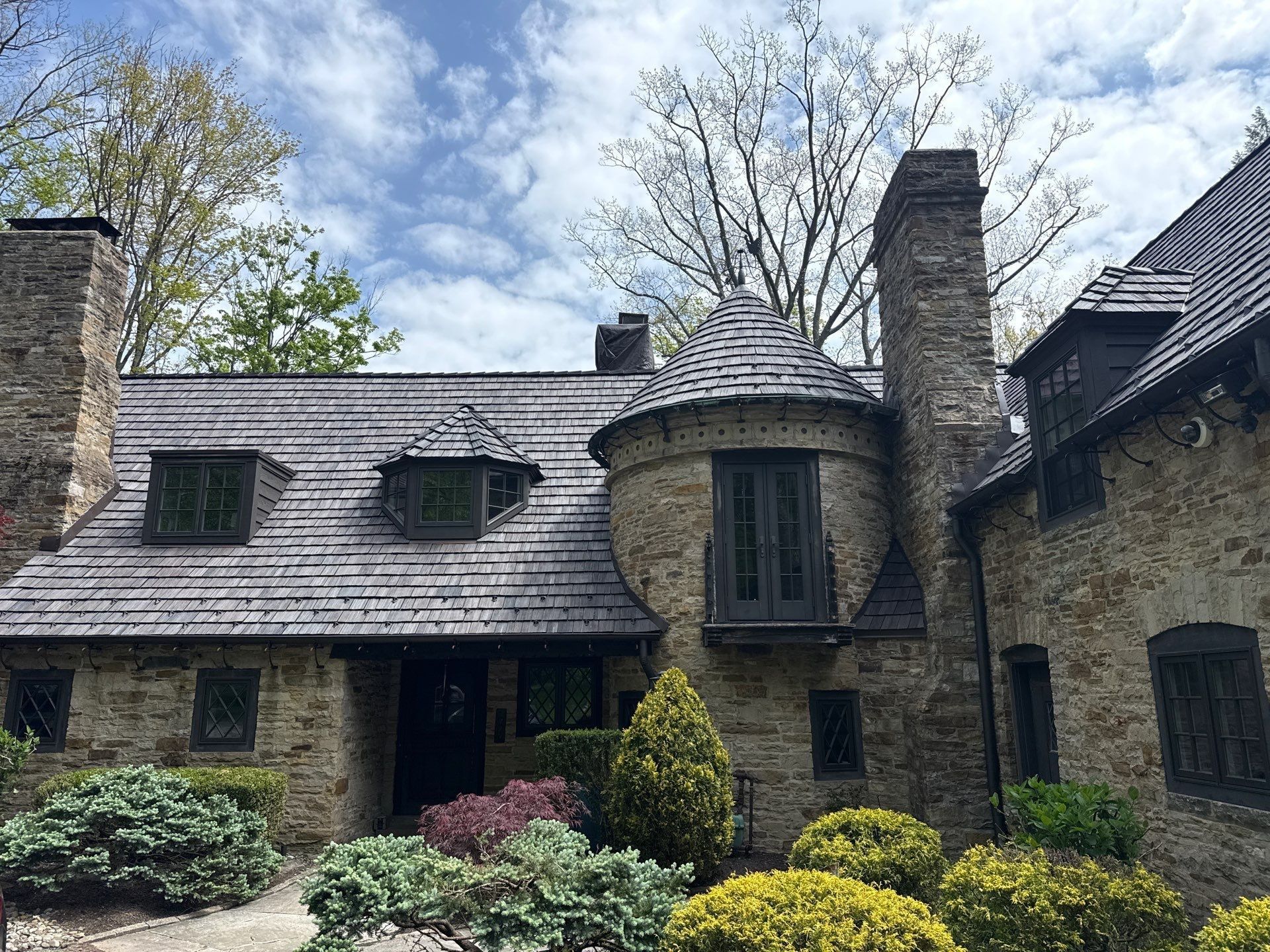 Stone cottage exterior with shingled roof, tall chimney, arched entry, and manicured shrubs under a cloudy sky