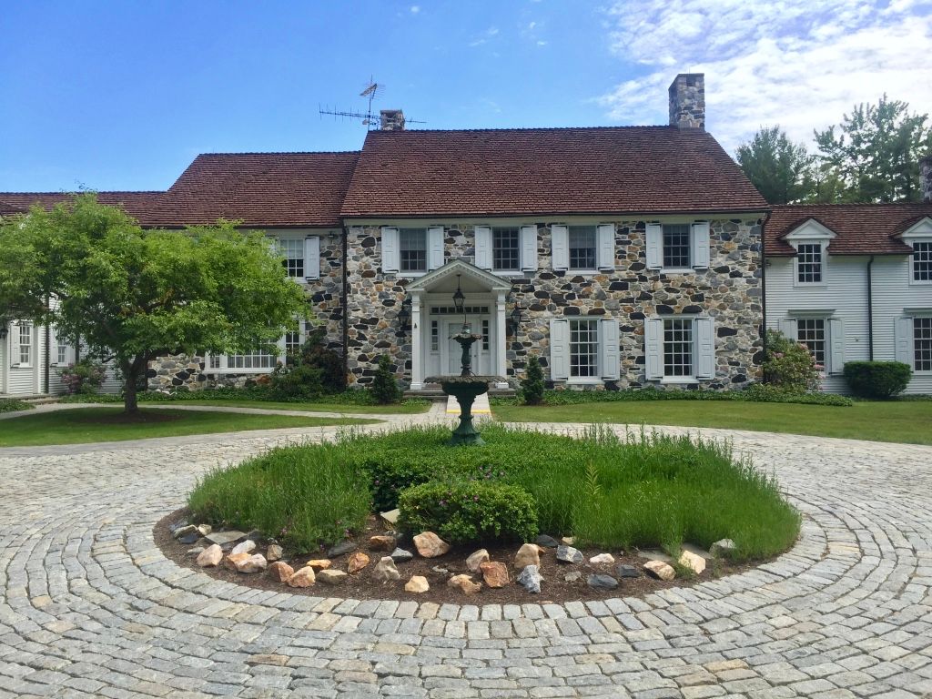 Historic stone house with red roof, white shutters, and circular cobblestone driveway around a central flowerbed