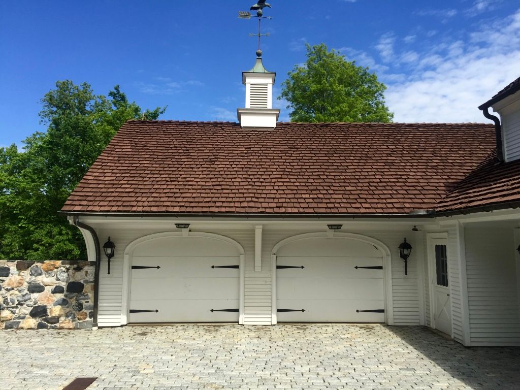 White double garage doors under a red shingled roof beside a stone wall and trees