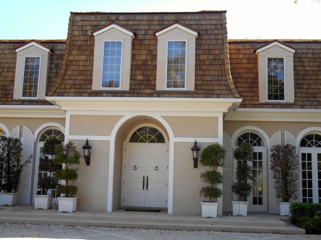 Front entrance of a beige house with white trim, arched doorway, dormer windows, and potted trees