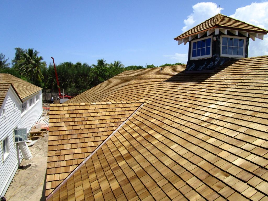 Large tan shingled roof with a small cupola under a clear blue sky, beside white houses and trees