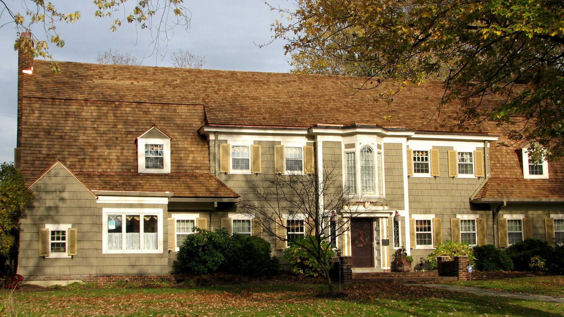 Two-story stone house with white-trimmed windows and a shingled roof in a wooded yard
