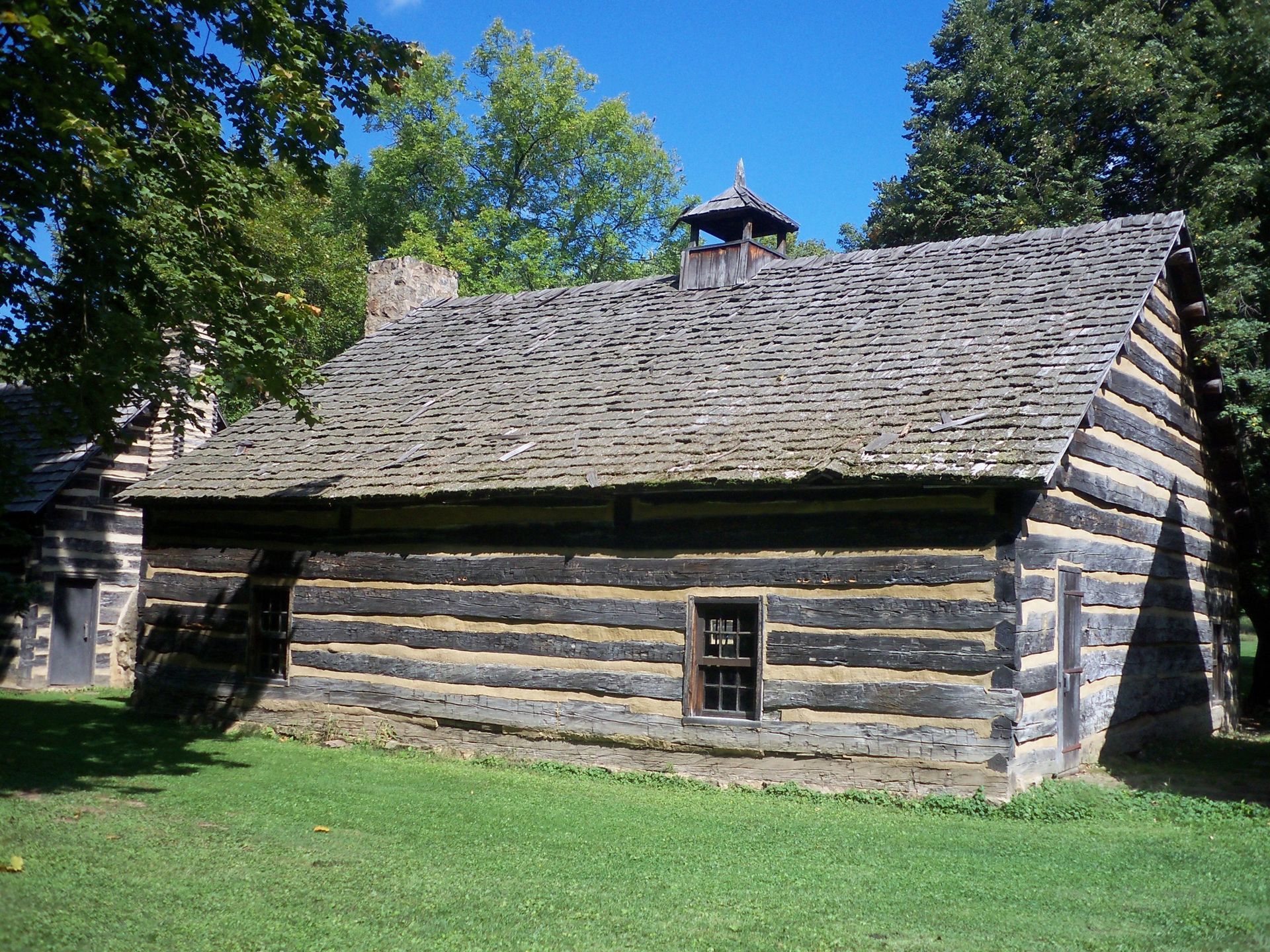 Log cabin with a steep shingled roof in a grassy, tree-lined setting