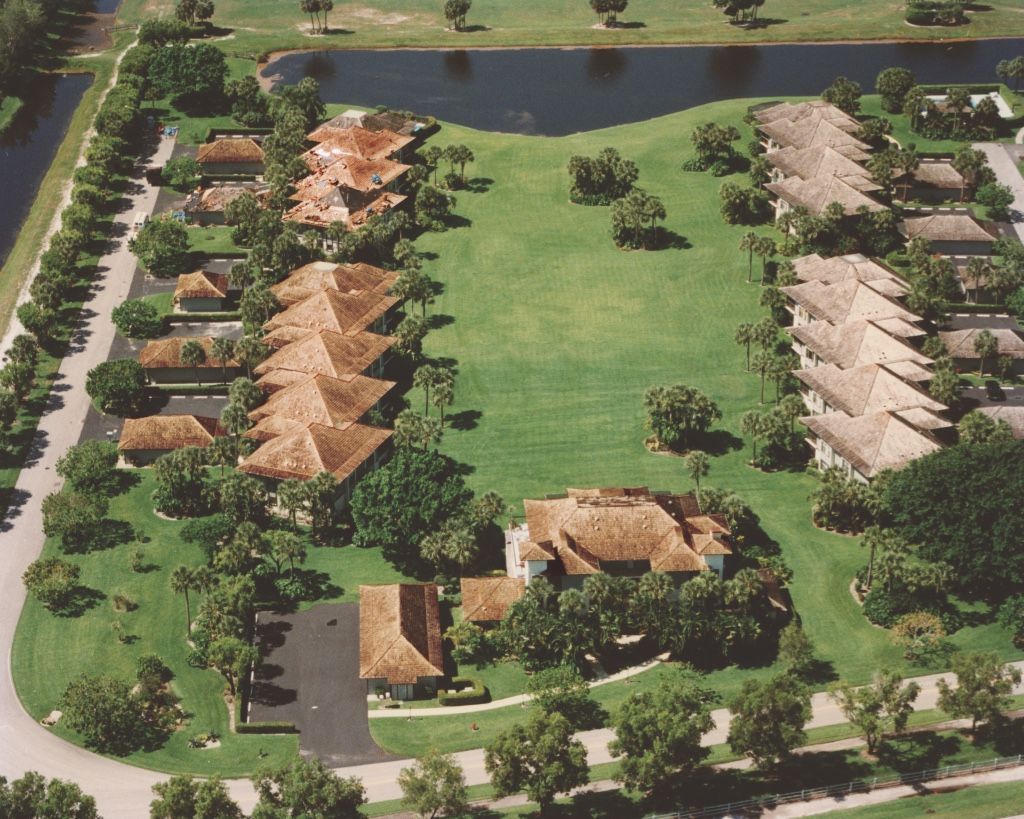 Aerial view of a resort with rows of tan-roofed huts, palm trees, and a central grassy courtyard by water.