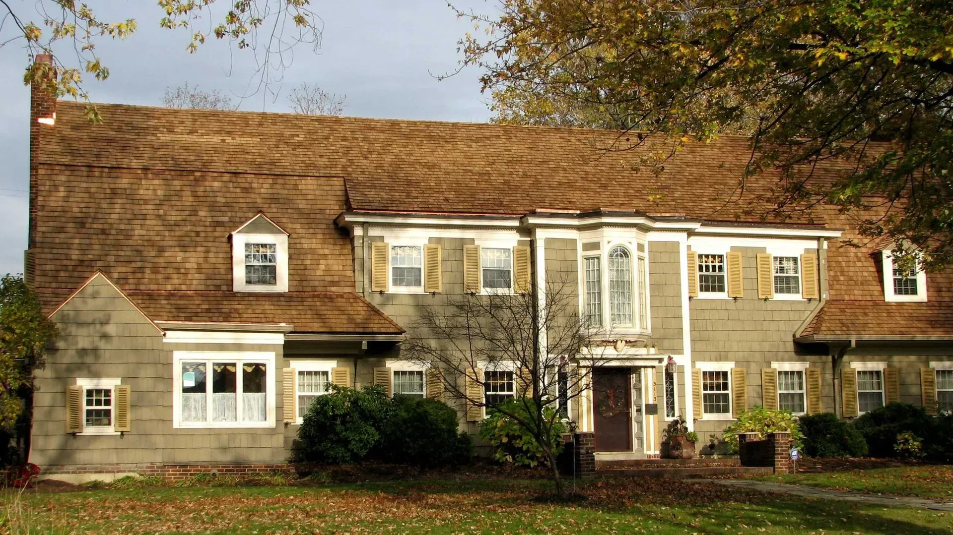 Stone house with brown shingled roof, white windows, and autumn trees in front