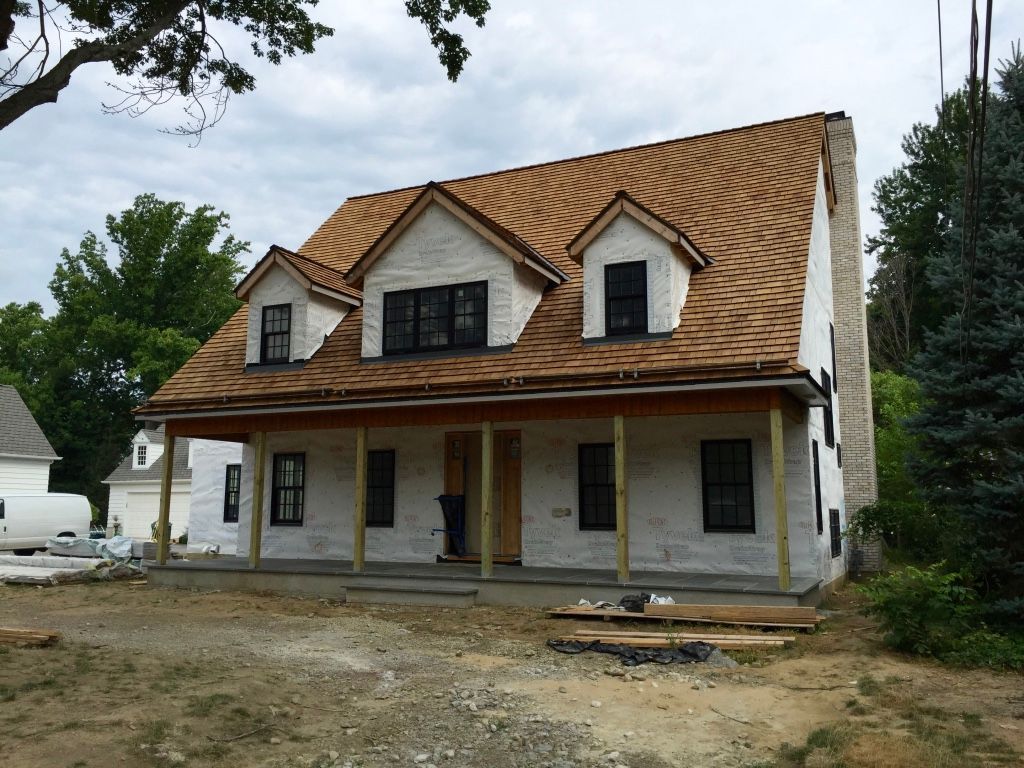 Two-story house under construction with white siding, tan roof, and front porch in a wooded lot