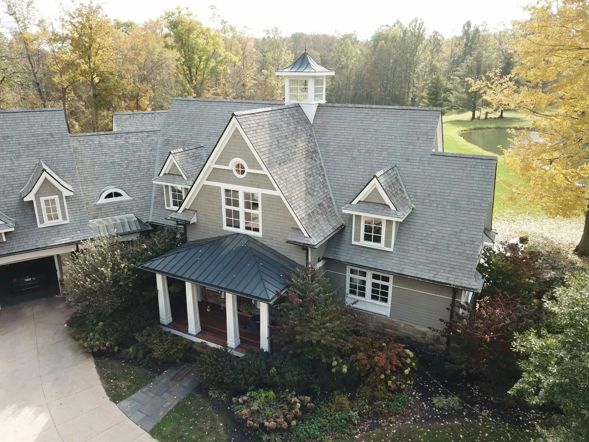 Aerial view of a gray-shingled house with a front porch, dormer windows, and surrounding trees.