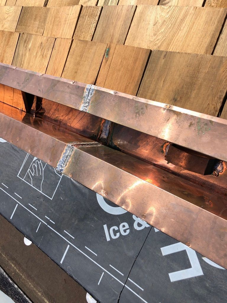 Close-up of a roof edge with copper flashing over black underlayment and tan shingles.