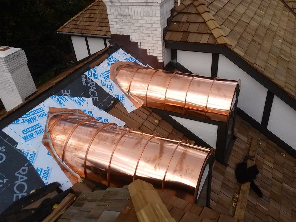 Two copper roof vents on a partially shingled rooftop beside a brick chimney and dormer windows.