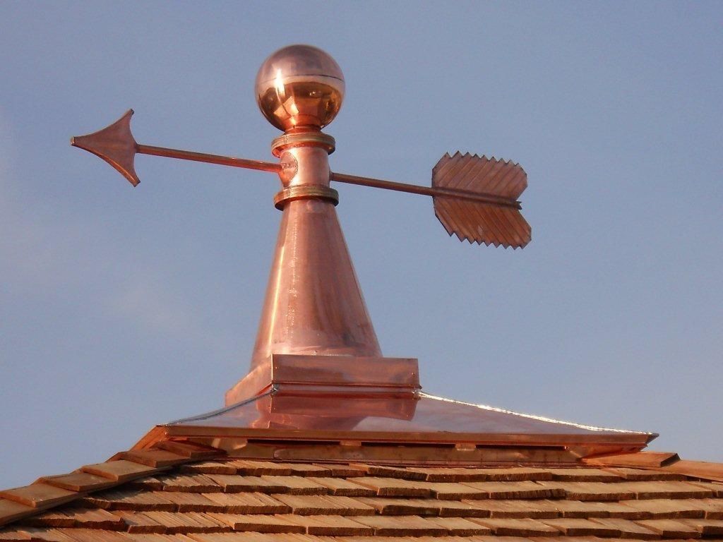 Copper weather vane on a roof with an arrow and feather-shaped finial against the sky