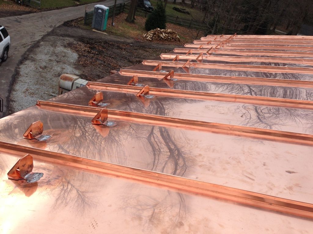 Copper-colored metal roof with several parallel ribs and clips, viewed from above near a driveway and trees