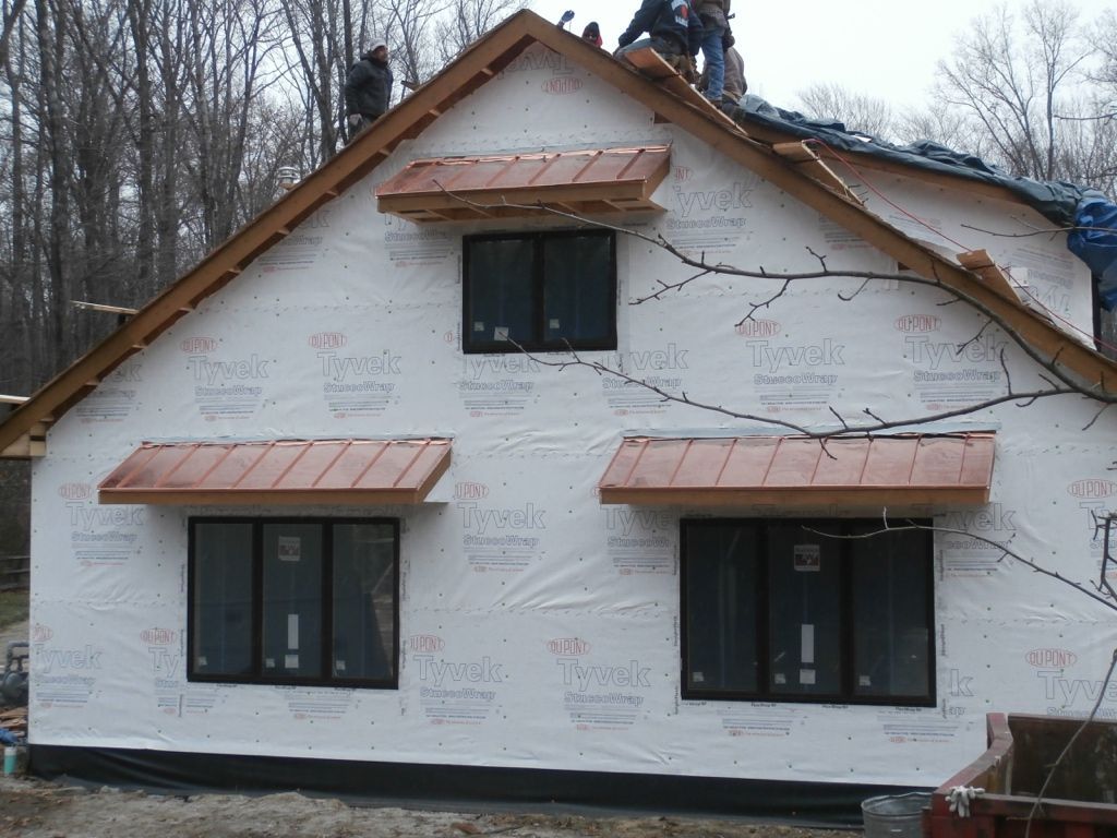 House under construction with white wrap, black windows, and copper awnings in a wooded area