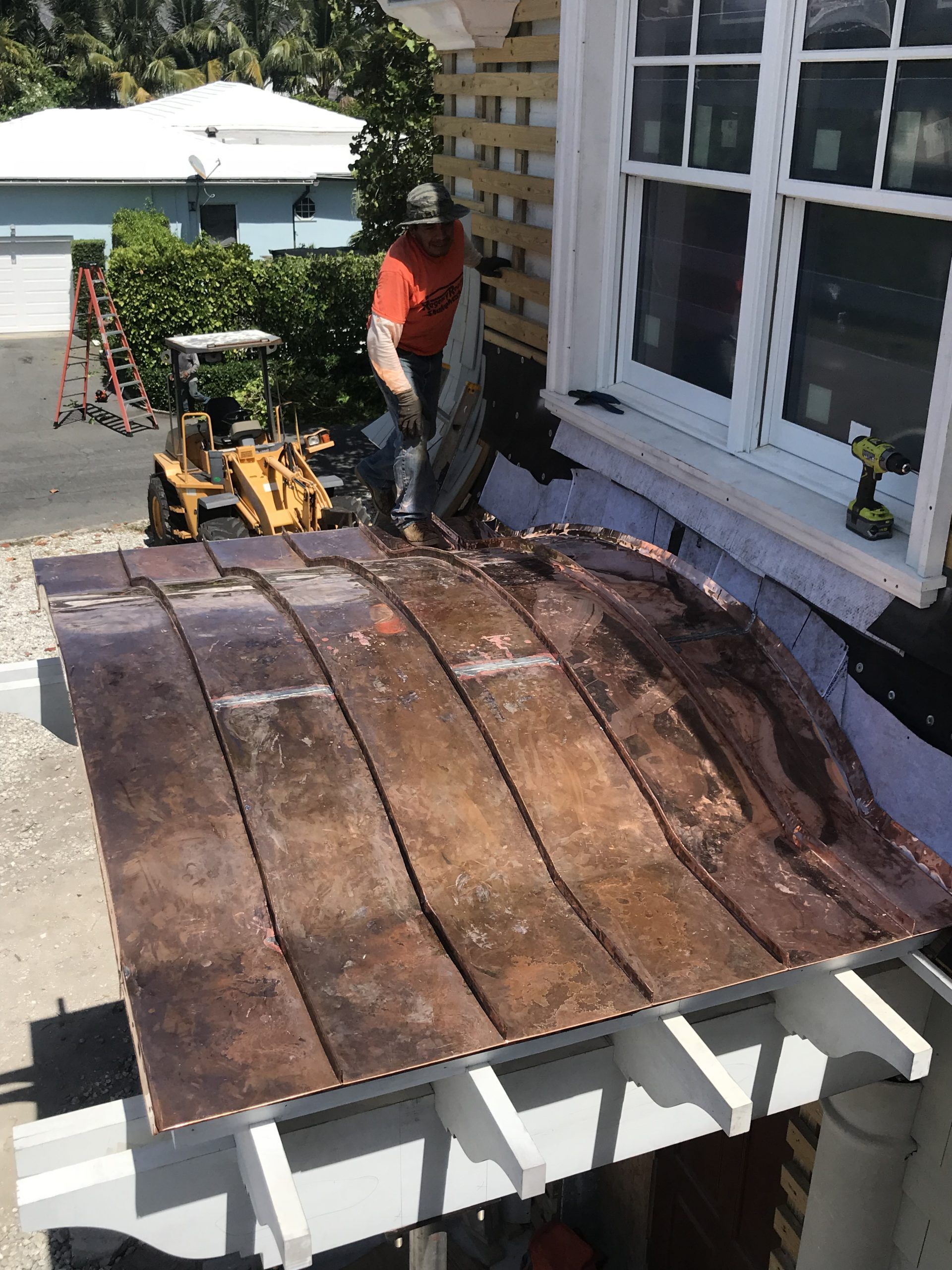 Worker repairing a weathered roof beside a white house, with tools and materials on a porch below.