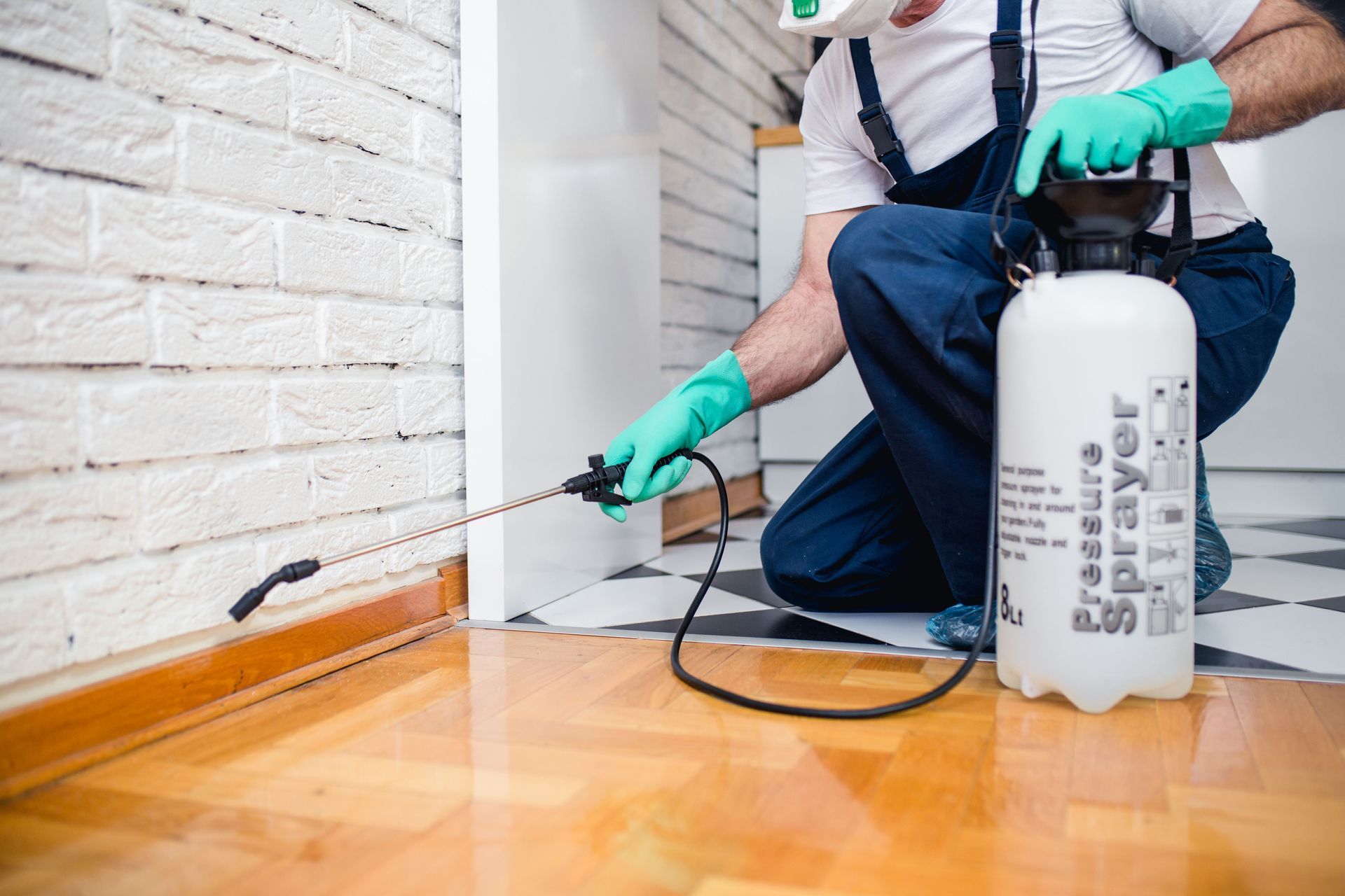 Pest control worker spraying along a baseboard in a room with wooden floor and white brick walls