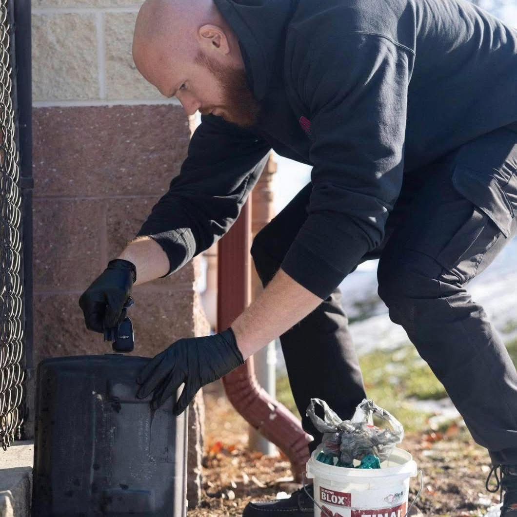 Man in black gloves cleaning a black container outdoors beside a wall and bucket