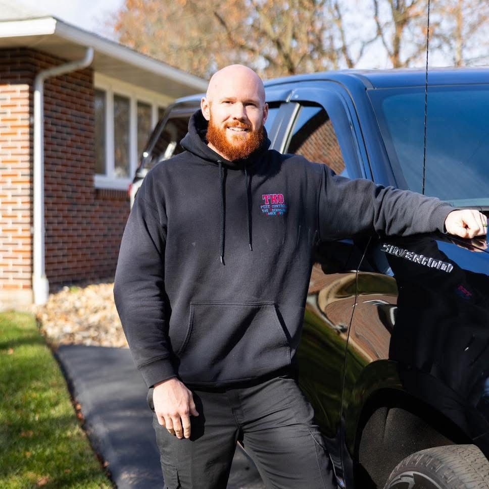 Man in a black hoodie leaning on a black truck beside a brick house on a sunny day