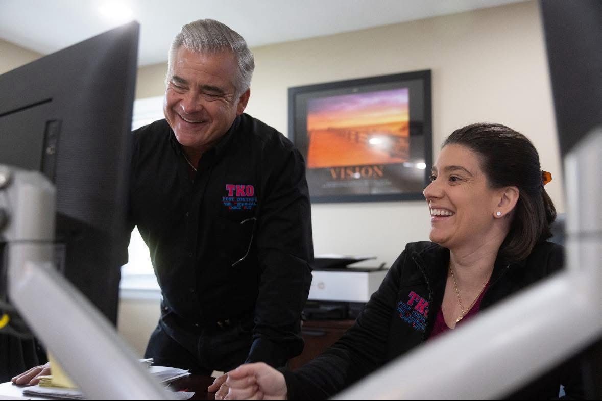 Two coworkers smiling at a computer in an office, one standing and one seated.