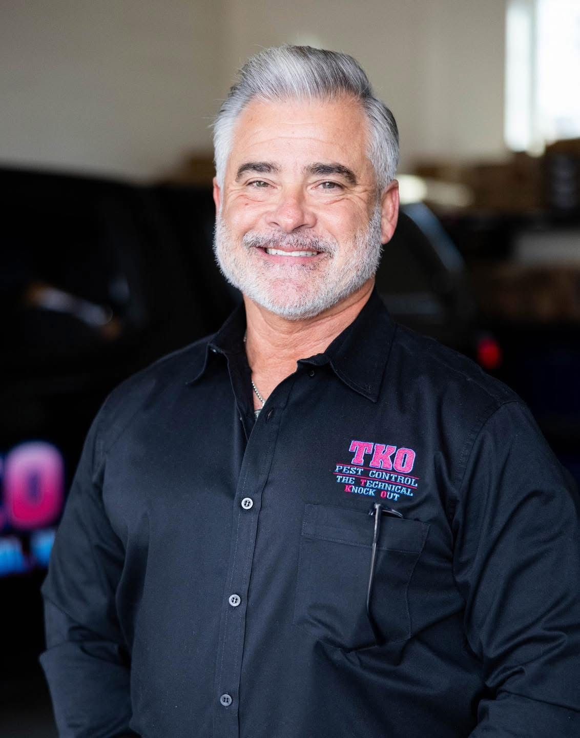 Smiling man in a black button-up shirt with pink embroidered logo, standing indoors.